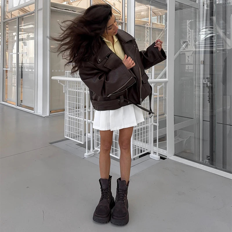 Woman in motion wearing the dark brown oversized moto jacket open over a white mini skirt and chunky boots, showing the dropped-shoulder silhouette