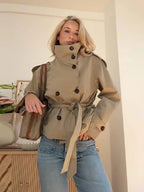 Women's cropped khaki trench jacket with double-breasted buttons, high stand collar and self-tie belt, styled with blue jeans – front three-quarter view
